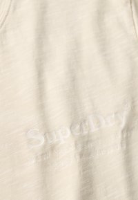 Textil texturizado de un beige claro con el logo de "SuperDry" y la dirección de Nueva York en blanco en relieve. Presenta un sutil patrón de rayas horizontales.