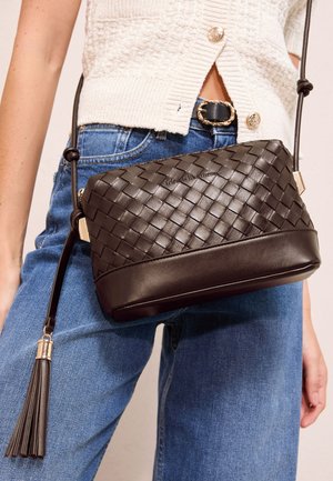 Brun flettet læder crossbody taske med kvastdetalje båret med blå jeans og en cremefarvet strikket knappet top.