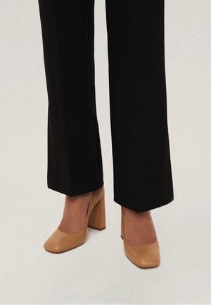 Beige High Heels mit quadratischer Zehenform, glänzender Oberfläche und einem blockförmigen Absatz, kombiniert mit schwarzen weiten Hosen.