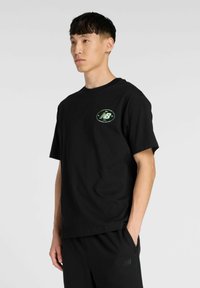 T-shirt en coton noir à manches courtes, avec un col rond et un logo ovale vert sur le côté gauche de la poitrine. Design simple sans motifs.