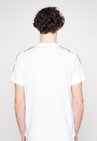 Een persoon met krullend haar, gezien van achteren, draagt een eenvoudig wit T-shirt met korte mouwen met zwarte strepen op de schouders, tegen een witte achtergrond.