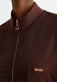 Brązowa kurtka zapinana na zamek z wysokim kołnierzem, wykonana z teksturowanej tkaniny. Posiada dekoracyjne logo w różowym złocie w pobliżu zamka.