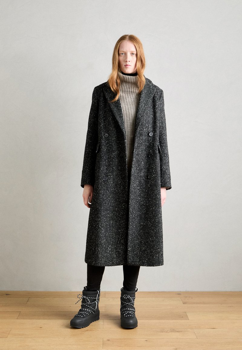 Długa czarno-biała płaszczowa peleryna w kropki z dużymi guzikami, noszona na szarym, prążkowanym golfie. Czarne, sznurowane botki za kostkę z teksturowaną podeszwą.