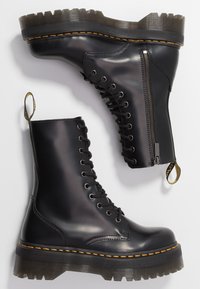 Botas de tobillo de cuero negro con suela texturizada, cierre de cordones en la parte delantera, cremallera lateral y acentos de costura amarilla. Diseño robusto con lengüeta para facilitar el calzado.