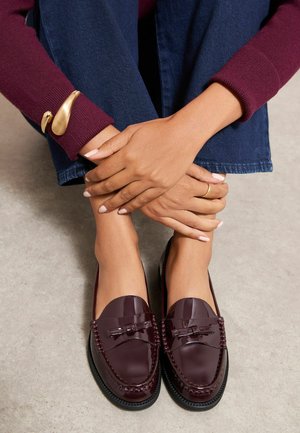 Mains avec une bague en or et un bracelet reposant sur des jambes croisées portant un jean bleu et des mocassins bordeaux brillants sur un sol clair.