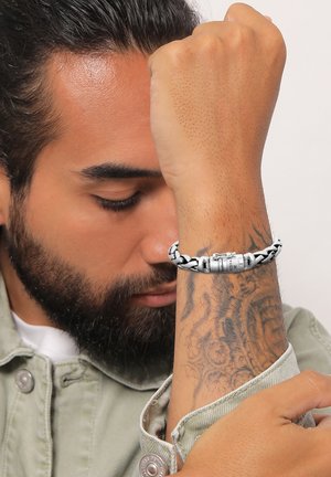 Flettet sølvarmbånd med en metallisk clasp, der har en tekstureret overflade og sorte accenter, båret på en underarm med indviklede tatoveringer.