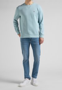 Felpa azzurro chiaro con scollo girocollo, polsini e orlo a coste, abbinata a jeans azzurri e sneakers bianche. Logo discreto sul petto.