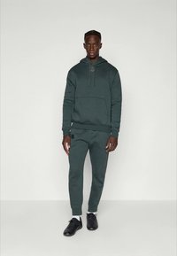 Felpa con cappuccio verde scuro e pantaloni jogger coordinati, realizzati in materiale morbido, con tasca frontale e dettaglio del logo. Indossati con scarpe da ginnastica nere.