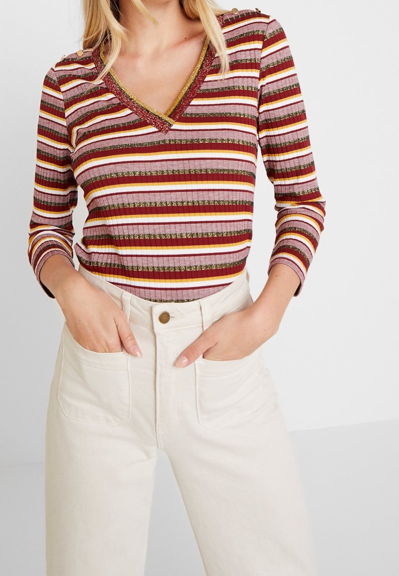 Femme portant un top ajusté à manches longues rayé avec des accents dorés et un pantalon taille haute écru avec poches avant, mains dans les poches.
