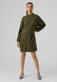 Abito in maglia di colore verde scuro, con collo alto, maniche lunghe e un fit rilassato. Abbinato a tacchi a punta di colore azzurro chiaro.