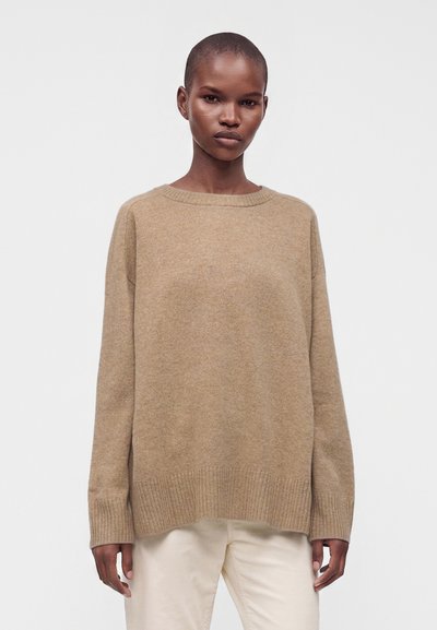 Jeune personne aux cheveux courts portant un pull beige ample et un pantalon clair sur un fond uni clair.