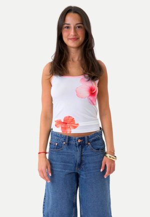 Jeune femme aux longs cheveux foncés porte un débardeur blanc avec des imprimés floraux roses et un jean bleu décontracté, debout devant un fond blanc.
