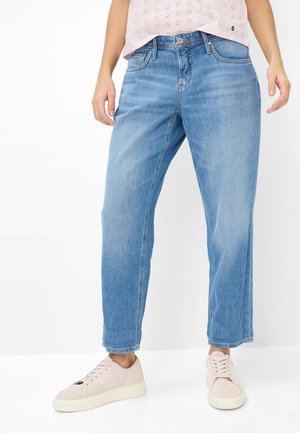 Frau trägt hellblaue Jeans mit geradem Bein, blassrosa Spitzenoberteil und hellrosa Sneaker, steht vor einem weißen Hintergrund.