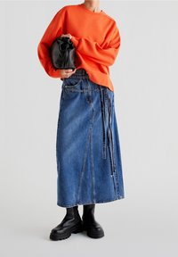 Persona vistiendo un sudadera naranja brillante, una larga falda de mezclilla azul con detalles de cinturón, botas negras gruesas y sosteniendo un bolso negro tipo pouch.