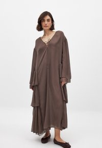 Lang bruin doorschijnend jurk met een v-hals, strikdetail, gelaagde tiered lagen en brede mouwen. Gecombineerd met donkerbruine platte schoenen.