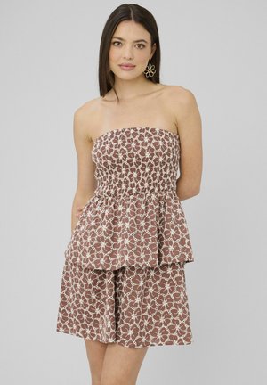 Femme aux cheveux raides et foncés portant une robe marron sans bretelles avec un motif en dentelle florale blanche et de grandes boucles d'oreilles en forme de fleur.