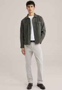 Donkergroene, knoopsluiting jas met twee borstzakken, gedragen over een witte t-shirt, gecombineerd met lichtgrijze broek en donkerbruine schoenen.