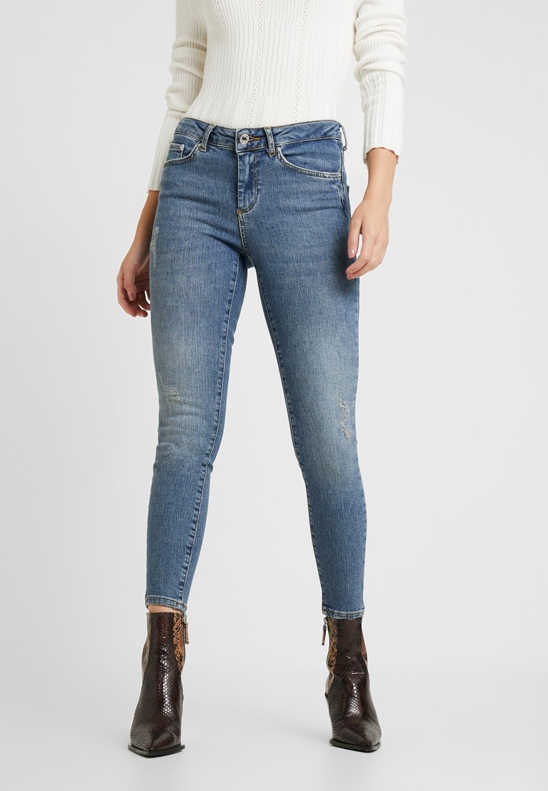 Person i blå skinny jeans med subtila slitage och bruna ankelboots med ormskinnsmönster, stående mot en neutral bakgrund.