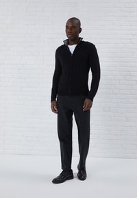Svart zip-up cardigan med en strukturerad stickning, med hög krage och ribbade ärmslut, kombinerad med svarta byxor och skor. Vit bakgrund.