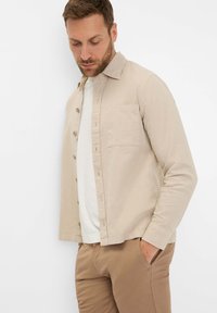 Light beige button-up shirt with a chest pocket, long sleeves, and a relaxed fit, layered over a white t-shirt and paired with brown pants.