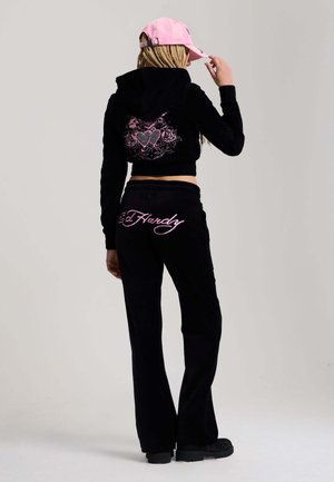 Osoba z plecionymi włosami, nosząca różową czapkę, czarną krótką bluzę z kapturem z różowym sercem i wzorem róż oraz czarne spodnie z różowym napisem „Ed Hardy”.