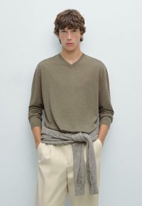 Maglione in lana color khaki a scollo a V abbinato a pantaloni beige chiari, con un maglione grigio in maglia annodato in vita; texture morbida e vestibilità rilassata.