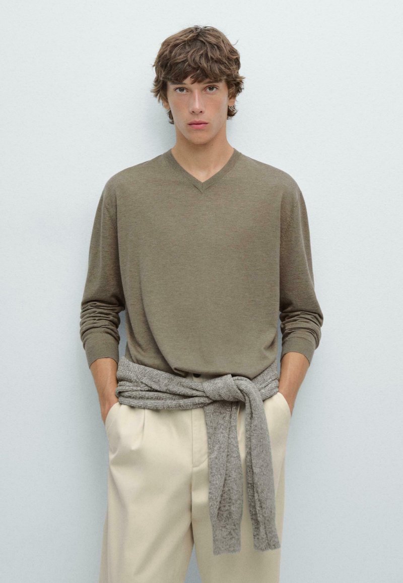 Maglione in lana color khaki a scollo a V abbinato a pantaloni beige chiari, con un maglione grigio in maglia annodato in vita; texture morbida e vestibilità rilassata.