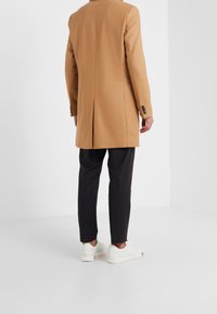 Kameluldsfrakke med lige snit, rygslids og sorte knapper, parret med mørke bukser og hvide sneakers på en ensfarvet baggrund.