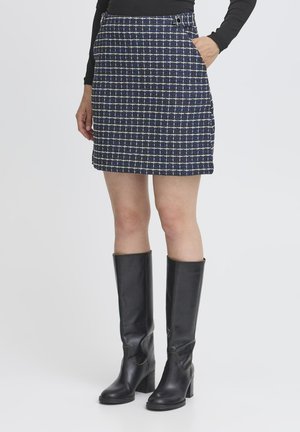 Mujer vistiendo una minifalda a cuadros en tonos azul marino y blanco, con una blusa de manga larga negra y botas de tacón altas negras, con la mano en el bolsillo.