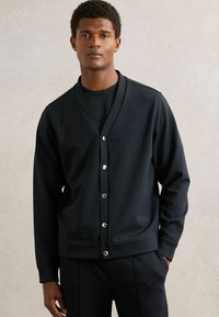 Cardigan nero in tessuto liscio, con scollatura a V, chiusura con bottoni e polsini a righe. Indossato sopra una camicia nera coordinata.