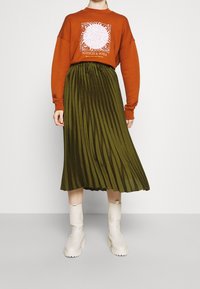 Sudadera naranja con logo gráfico blanco, combinada con una falda midi plisada verde y botines crema. Elementos de diseño simples y modernos.