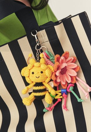 Háčkovaný žlto-čierny prívesok na kľúče v tvare včielky a ružový prívesok kvetiny pripevnené k čierno-bielemu pruhovanému taške na rameno.