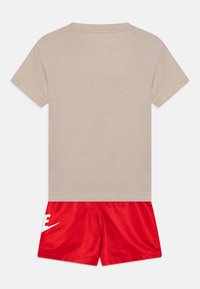 Bežové bavlnené tričko s krátkymi rukávmi a okrúhlym výstrihom, v kombinácii s červenými nylonovými šortkami s bielym logom Nike na ľavej strane.