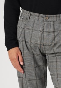 Pantaloni cu carouri gri, cu talie înaltă, având un detaliu cu nasture și buclă, țesătură texturată și accente maronii contrastante în model.