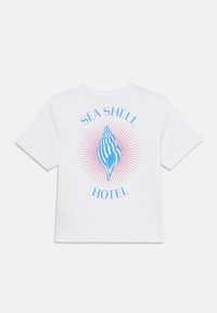 Camiseta de algodón blanca con un gráfico de concha azul y rayos rosas, y el texto "SEA SHELL HOTEL" alrededor en la parte posterior.