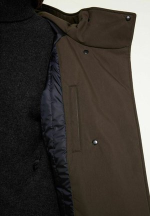 Olive green outer jacket with a smooth texture, featuring a black, quilted lining, snap buttons, and a side pocket. Black knit turtleneck underneath.