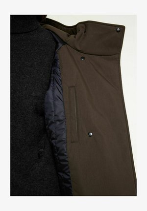 Olive green outer jacket with a smooth texture, featuring a black, quilted lining, snap buttons, and a side pocket. Black knit turtleneck underneath.
