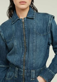 Denim jumpsuit in dark blue with long sleeves, front zip closure, and two chest pockets. Features a belted waist and subtle texture.