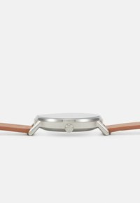 Rund klocka i rostfritt stål med en slät silverfärgad urtavla och ett rem av brunt läder. Har en sidokrona för inställningar.