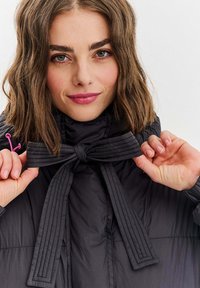 Czarna pikowana kurtka z kapturem, zapinana na pikowaną kokardę. Gładki materiał o delikatnym połysku z różowym akcentem w okolicy szyi.