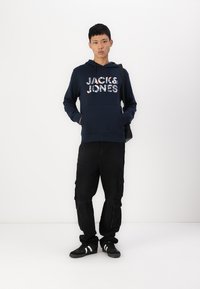 Marineblaue Kapuzenjacke mit "JACK & JONES" in einem mehrfarbigen Design bedruckt, kombiniert mit schwarzen Cargo-Hosen und schwarzen Sneakers.
