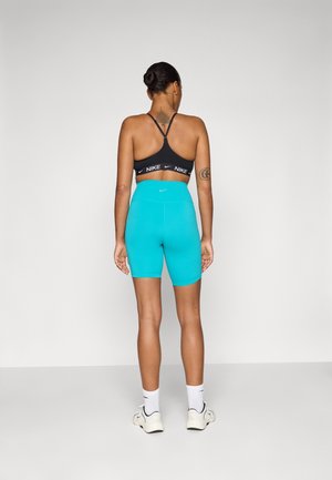 Vrouw staand met de rug naar de camera, draagt een zwarte Nike sport-bh, turquoise hoog getailleerde fietsbroek, witte sokken en witte sneakers op een effen achtergrond.