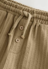 Beige textured lightweight trousers with a drawstring waistband, featuring two wooden buttons and subtle vertical stripes.