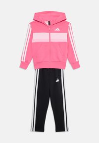 Veste à zip rose avec des rayures blanches, poches avant, et pantalon de survêtement noir avec trois rayures blanches et logo. Mélange de coton doux et athlétique.