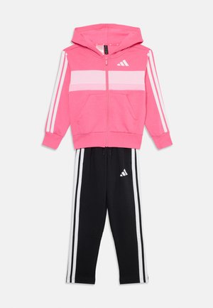 Veste à zip rose avec des rayures blanches, poches avant, et pantalon de survêtement noir avec trois rayures blanches et logo. Mélange de coton doux et athlétique.