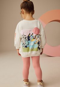 Sudadera blanca con un colorido gráfico de dibujo animado en la parte posterior, combinada con leggings de canalé rosas y zapatillas de color claro.