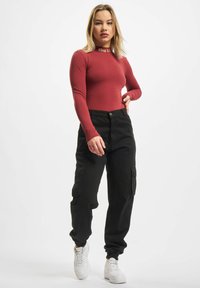 Rød langermet bodysuit med gull "Thug Life" tekst, parret med sorte cargo-bukser med sidelommer og ribbede mansjetter, komplementert av hvite joggesko.