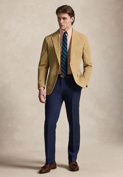 Homme portant un blazer beige, une chemise rayée orange, une cravate rayée verte et marine, un pantalon bleu marine et des mocassins marron, debout devant un fond uni.