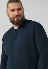 Dunkelblaues Jackett mit rundem Kragen, Knopfverschluss und seitlichen Taschen. Hergestellt aus glattem Stoff, mit einem strukturierten Muster und einem lockeren Schnitt.