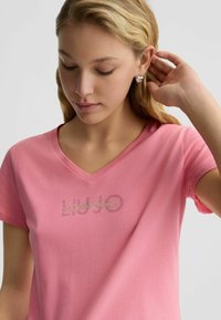 Rosa Baumwoll-T-Shirt mit V-Ausschnitt, das ein Strass-Logo auf der Vorderseite trägt. Der Stoff wirkt glatt, leicht und lässig im Stil.